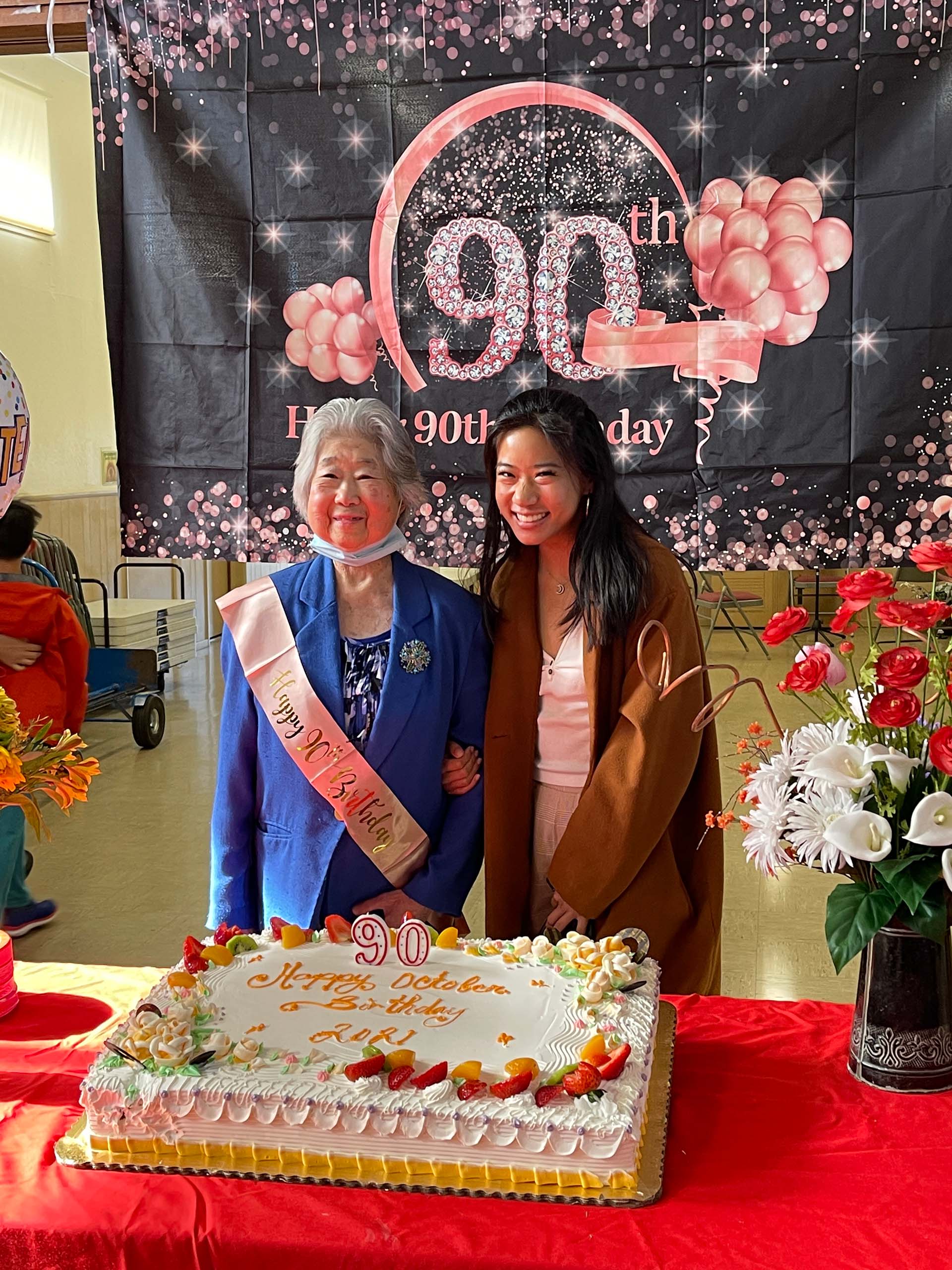 A young woman poses with her grandmother in front of a birthday during the grandmother's 90th birthday celebration. The older woman wears a pink sash that reads, "Happy 90th Birthday."