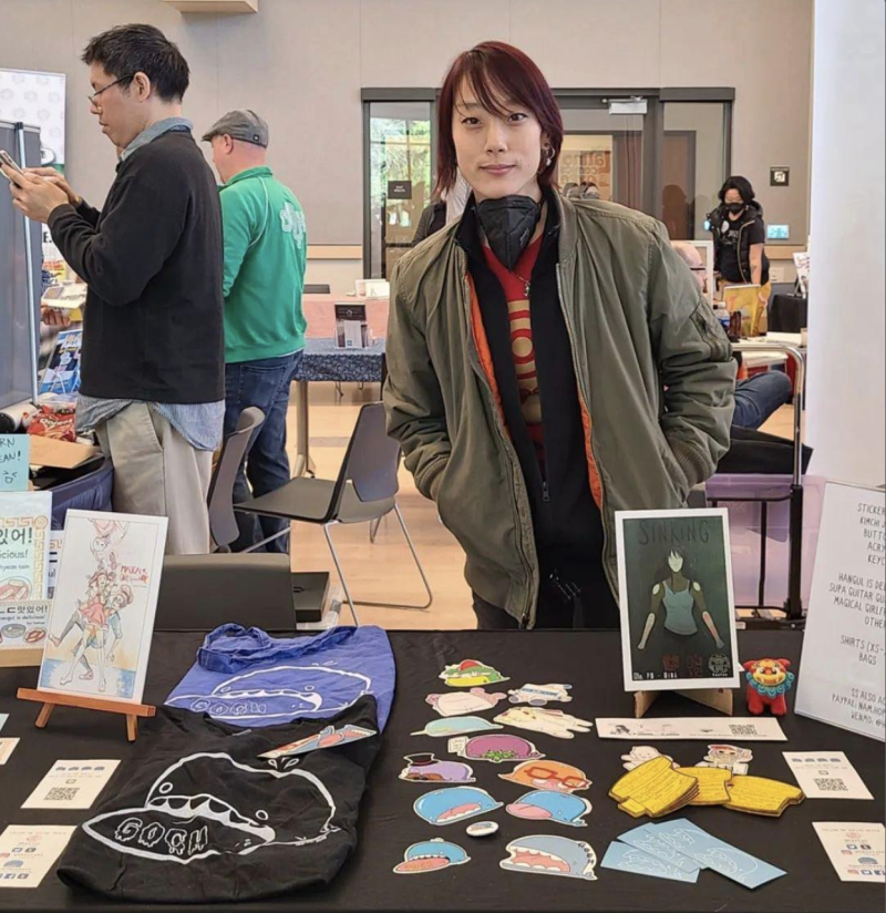 a young Asian woman poses behind a table full of zines and stickers and tshirts at a book fair