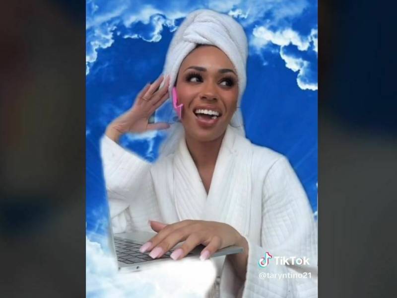 A beautiful young woman of color smiles and chats in front of a blue sky background. She is wearing a white bath robe and towel on her head.