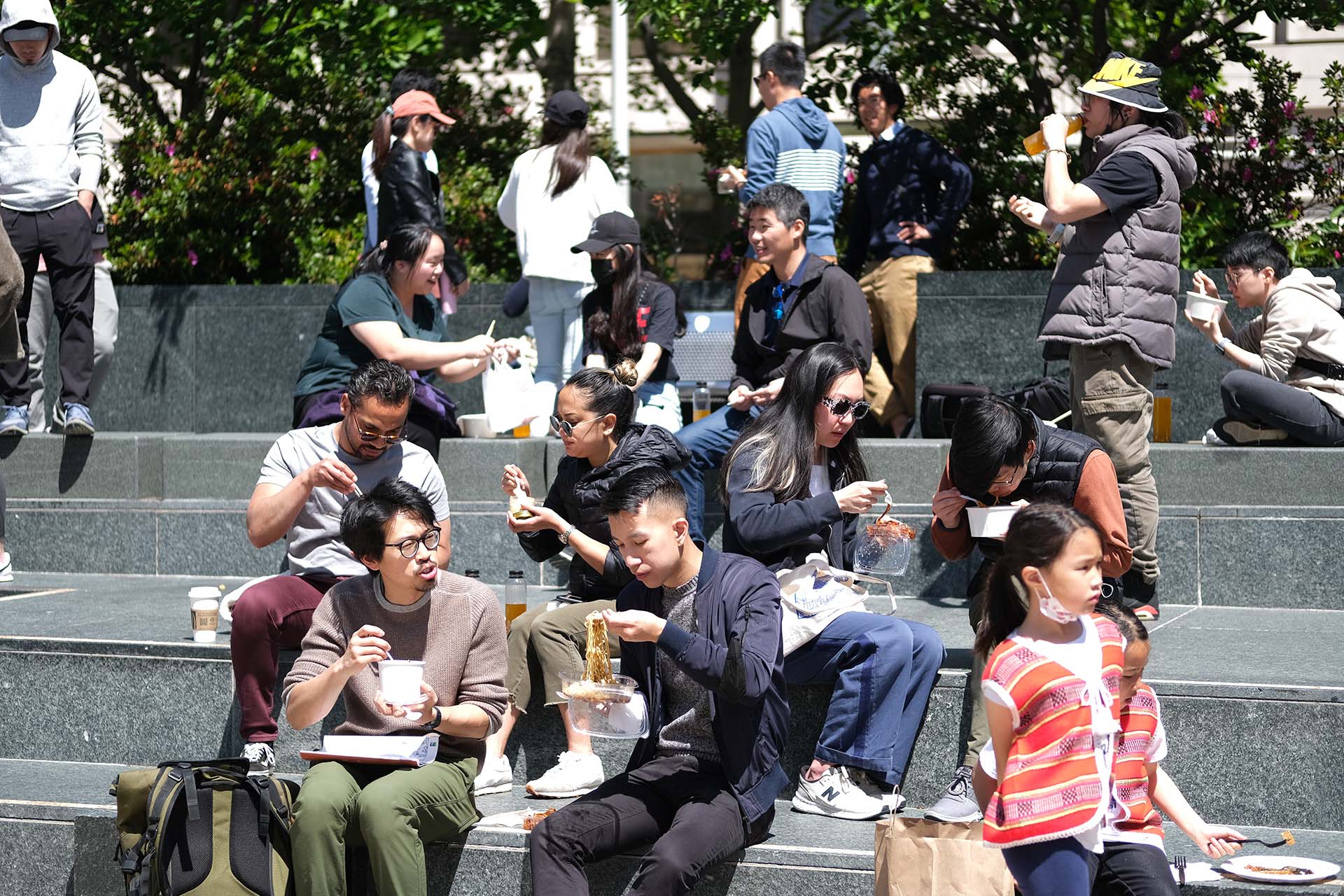 A crowd of people sit eating noodles from paper bowls on a set of stairs outside in Union Square, San Francisco.