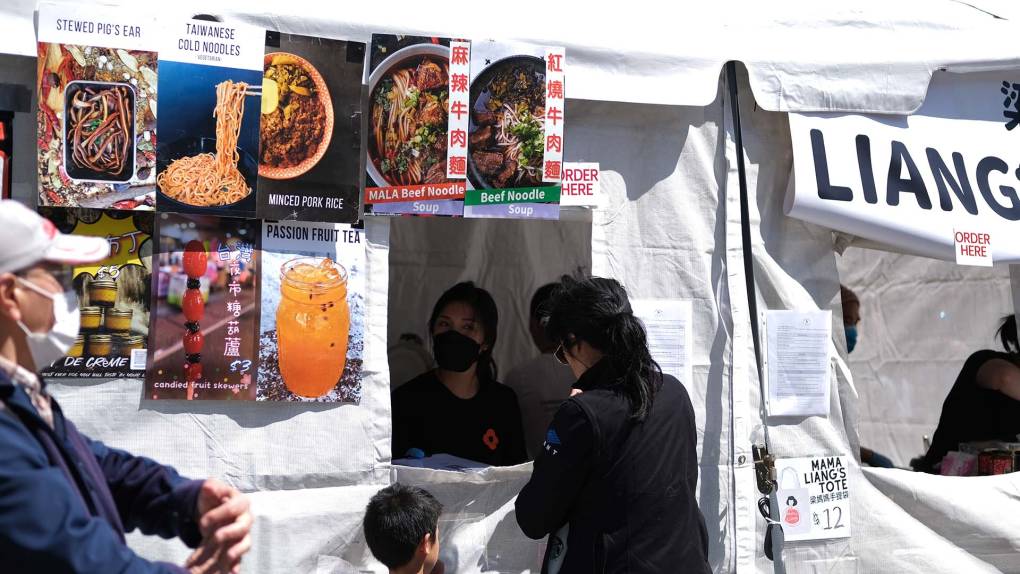 A customer orders food from the Liang's Village food tent, which has pictures of beef noodle soup and other Taiwanese dishes displayed.