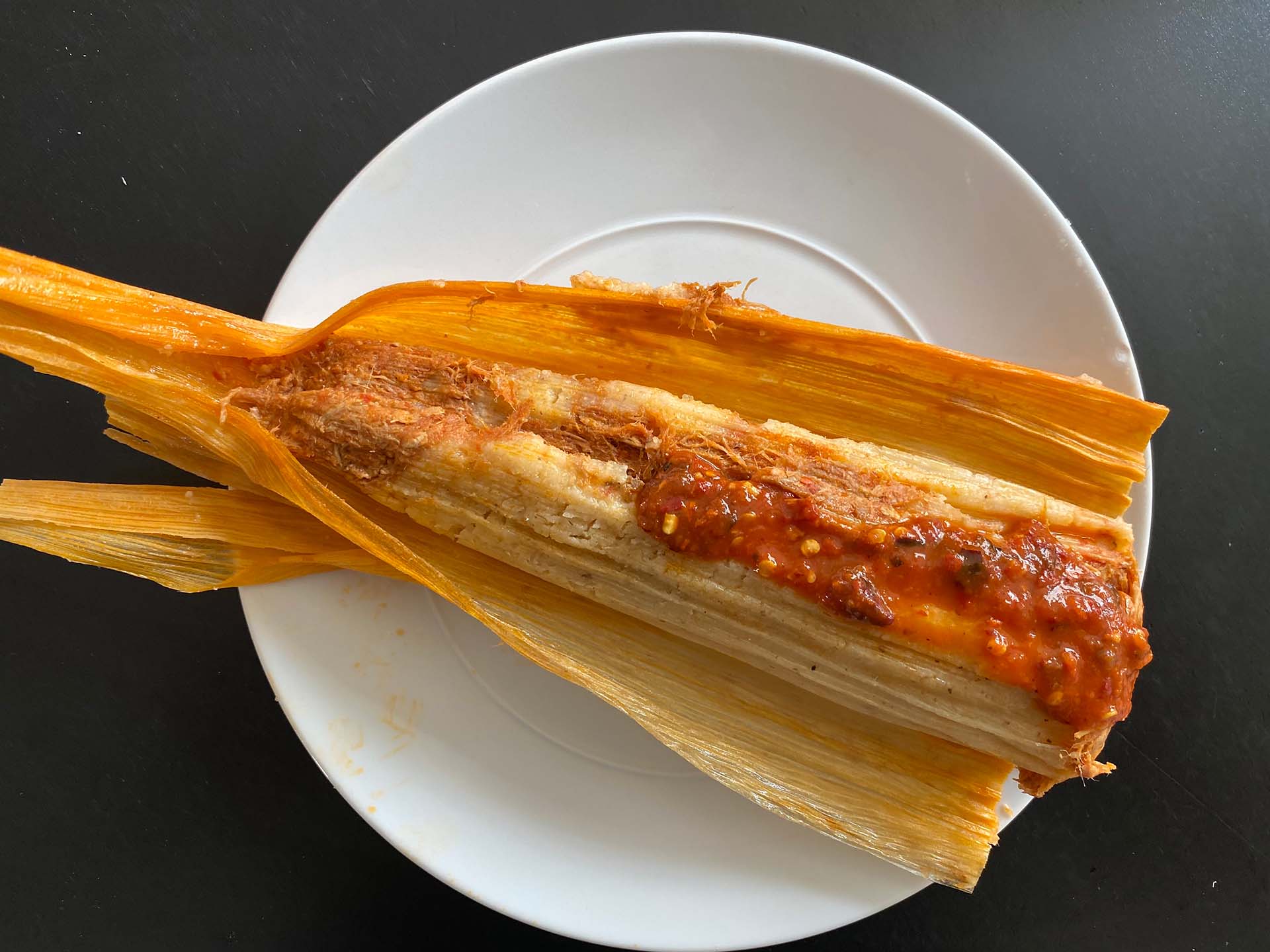 A tamal that has been opened. It has been topped with red salsa, and the cooked masa inside the corn husk is visible.