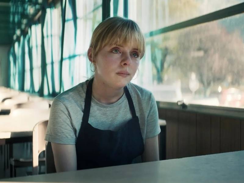 A young woman with blonde hair tied back sits at a diner table with a concerned expression on her face. She is wearing an apron.