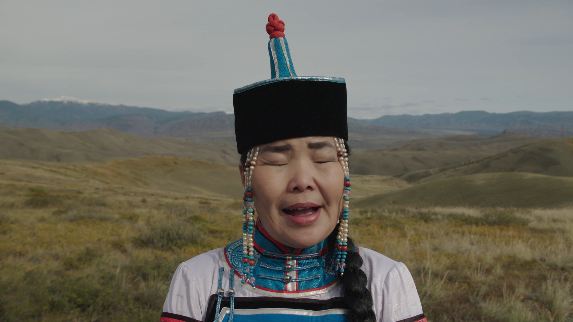 A Tuvan woman in traditional clothing sings with eyes closed, grassland in background