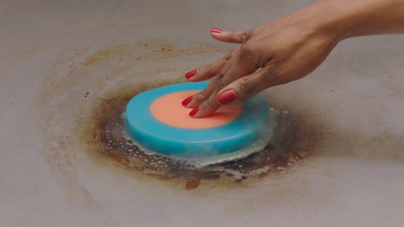 A hand presses a thick circular slice of blue and pink gelatin onto a hot surface
