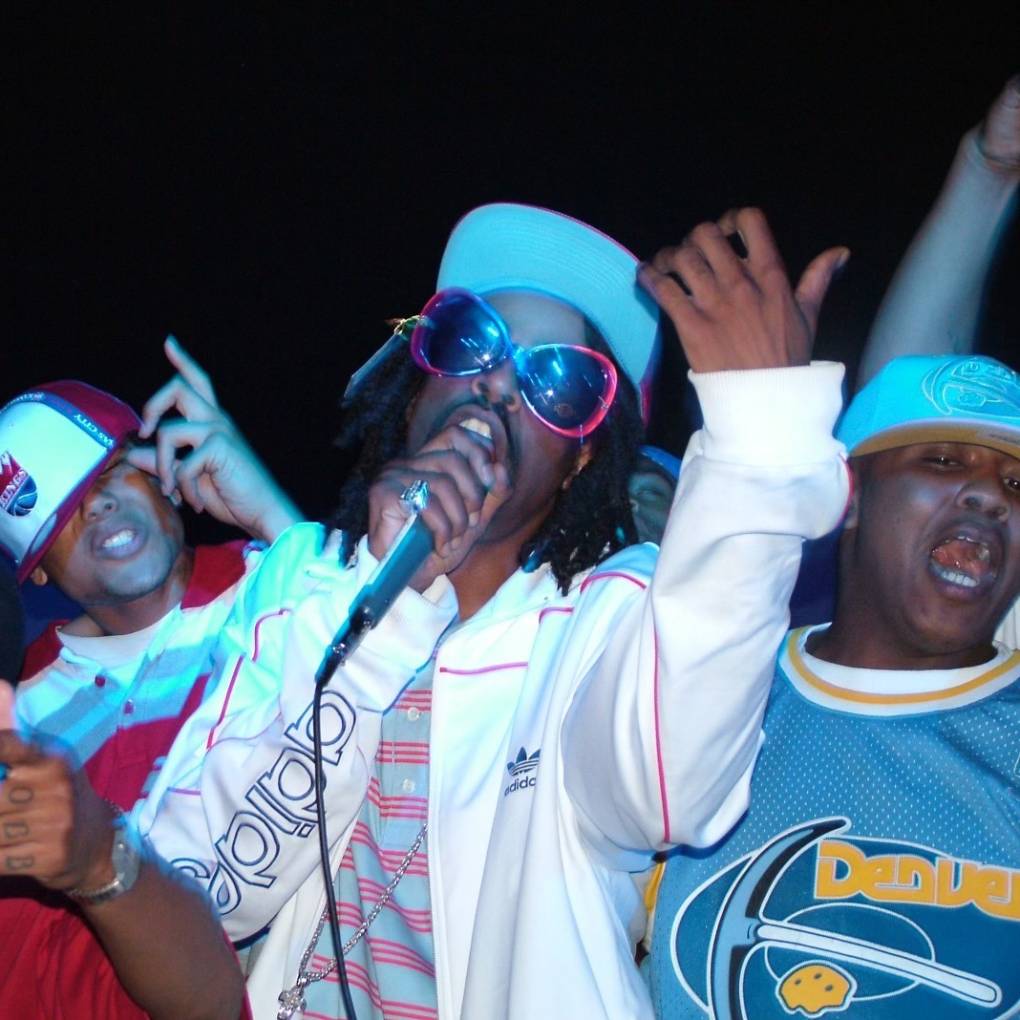 Mac Dre performs on stage in 2004, wearing giant sunglasses, a striped polo shirt and Adidas jacket. Men on stage are going dumb, dancing and singing along to the lyrics.