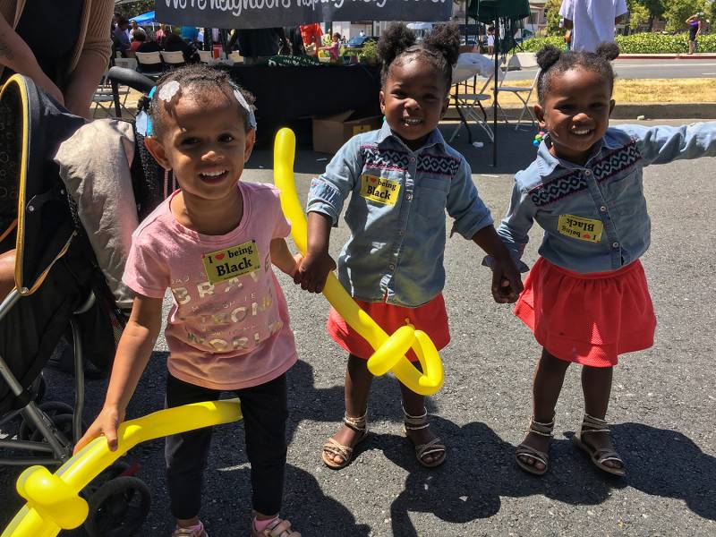 three adorable little Black girls hold balloon animals and wear stickers that read 'I heart being Black' at a festival