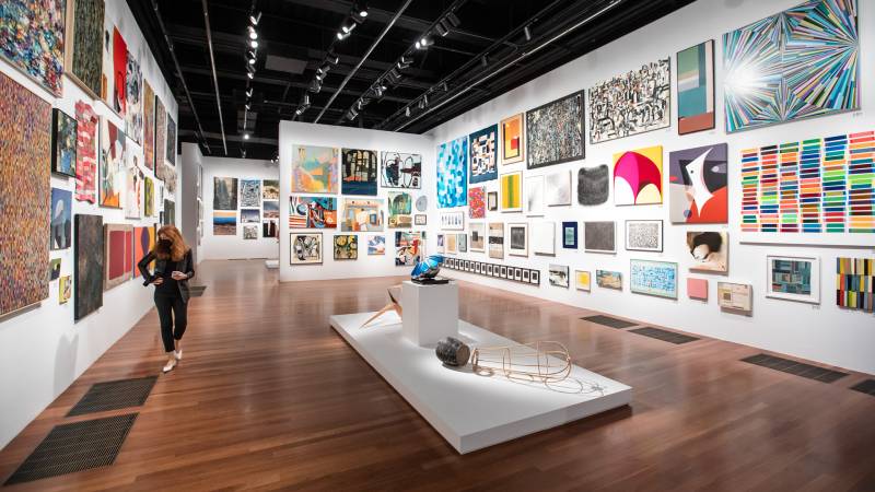 Wide view of salon-style hang of white walled gallery with hundreds of colorful paintings and artworks alongside each other.