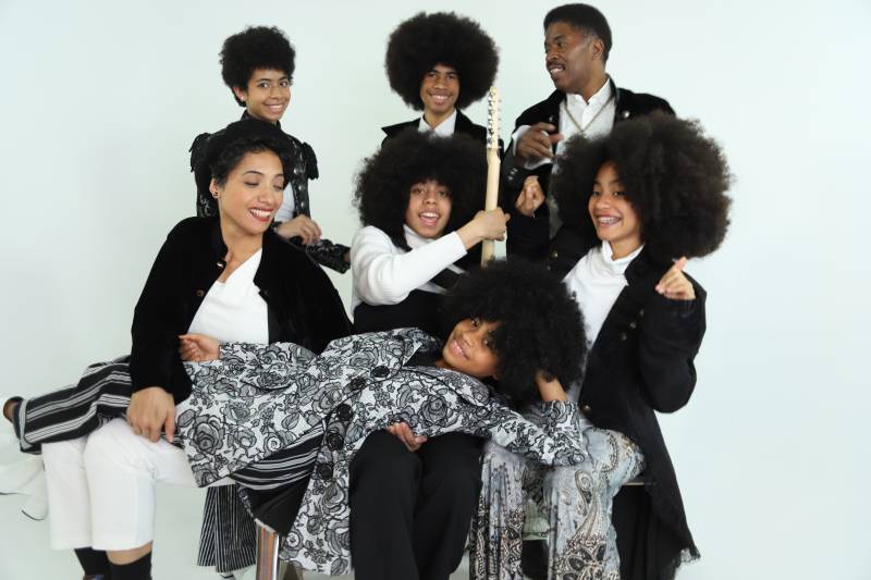 a musical group of seven people dressed in black and white, most of them with Afros, pose while holding instruments and smiling