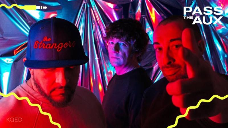 three musicians stand in a darkened room with neon lighting