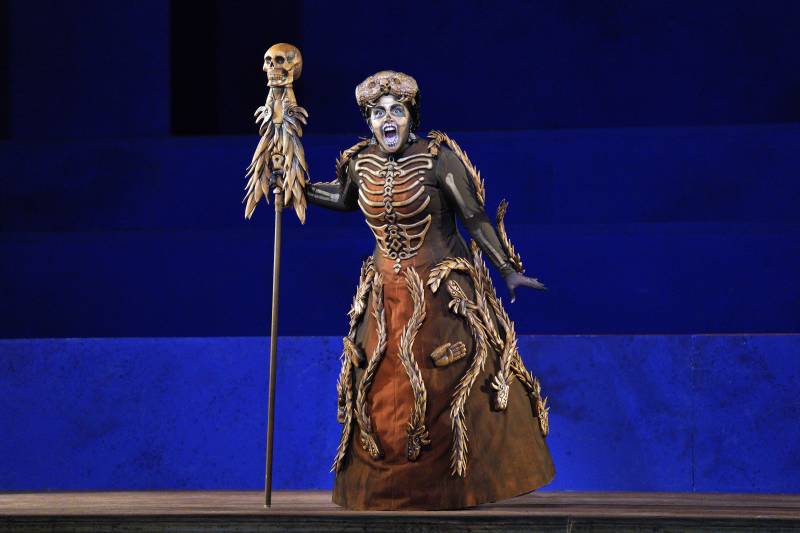 a person in an underworld-themed dress/skeleton costume holds a staff with a skeleton on top on a blue stage in a play