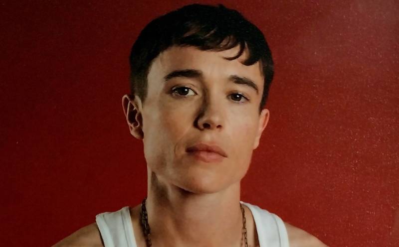 A transgender man sits, expression plain, in front of a red background. He is wearing a white tank.