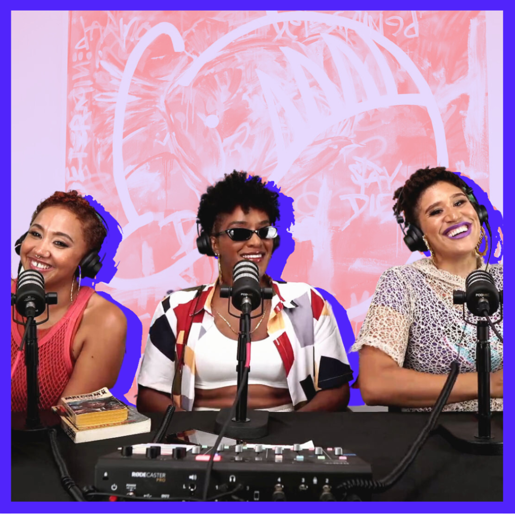Three Black women in colorful clothing sit in front of microphones while laughing.