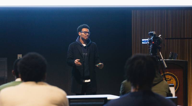 a young Black man in glasses and a black hoodie delivers a lecture