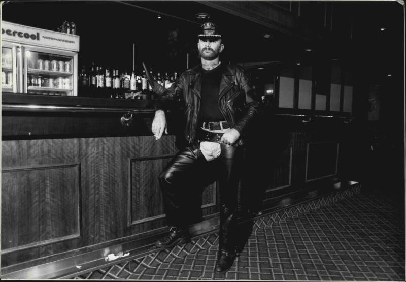 A man in leather chaps, jacket and hat, with black t-shirt on, stands assertively next to a wooden bar.