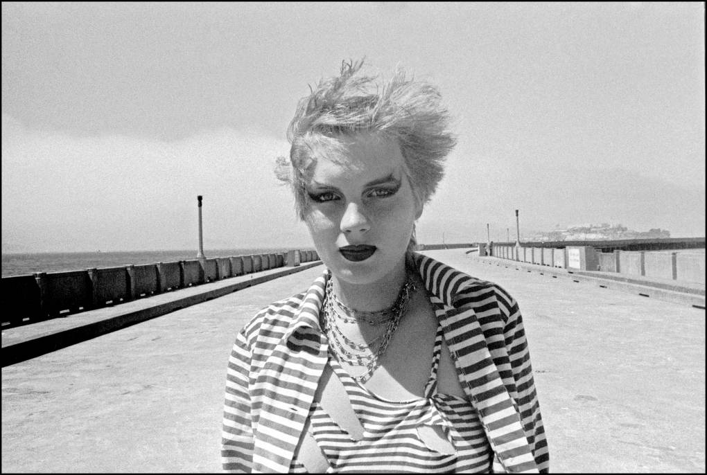 A white woman wearing black lipstick and heavy eyeliner stands windswept on the shoreline. Her bleached blond hair is short and she is wearing a safety pin necklace and ripped striped shirt and jacket.
