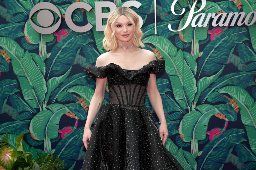 A very petite blonde woman wearing an off the shoulder black gown stands on a red carpet against a green backdrop.
