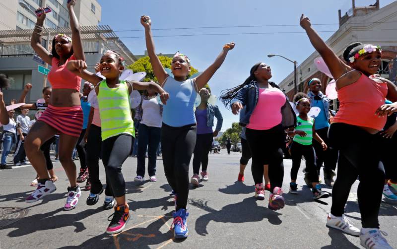 a group of joyous young Black girls in colorful shirts dance in the street as part of a parade 