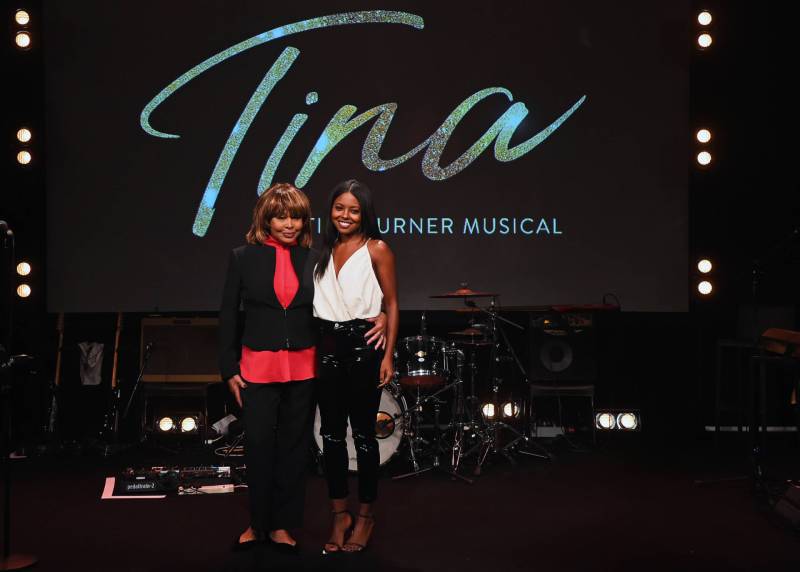 A senior Black woman wearing red blouse and black pants warmly embraces a younger, smiling Black woman wearing white top and black pants. They are standing on a stage with the word Tina lit up behind them.
