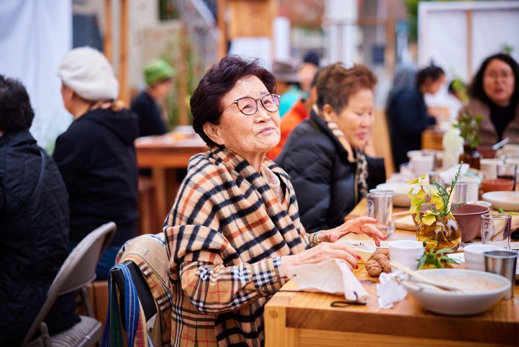 A elderly Korean woman in glasses looks on with a wistful expression on her face.