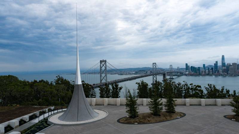 A gray and silver sculpture sits on a hill with a bridge, water and buildings in the background.