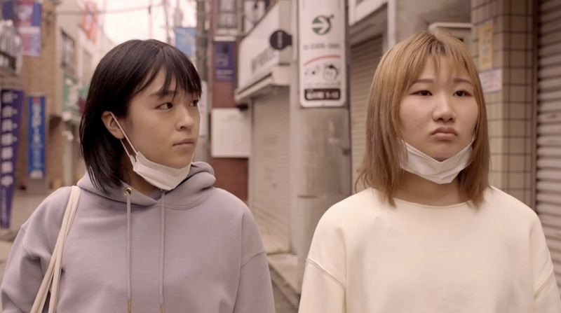 two young Asian women with masks around their chins walk on the street in a still from a film