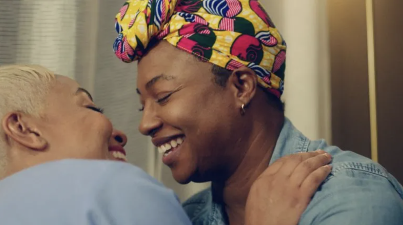 two Black women laugh in an intimate embrace; both wear light blue shirts and one wears a colorful head wrap