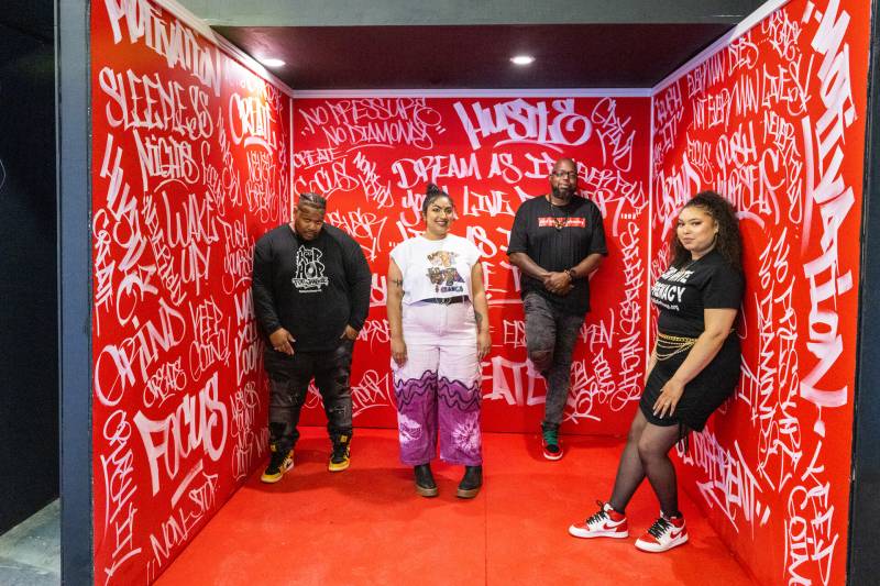 The four Hip-Hop for Change leaders, two men and two women, stand in front of a red studio backdrop covered in graffiti.