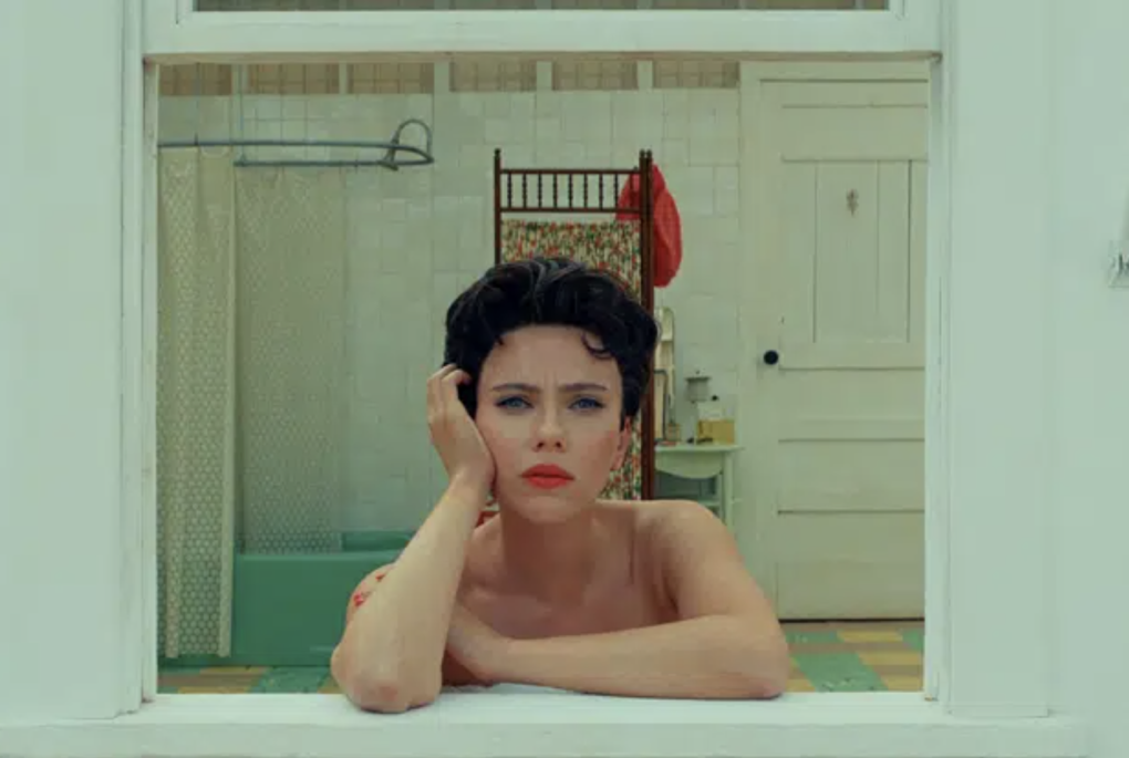 A woman with short brown hair curled on top of her head sits in an open window, looking forlorn and leaning on her right hand. She is wearing striking red lipstick.