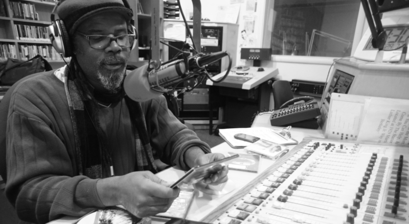 Greg Bridges is photographed in black and white as he sits behind a microphone and soundboard at KPFA studios in Berkeley, while hosting his jazz radio show. He is holding a cd case and reading the track list. 
