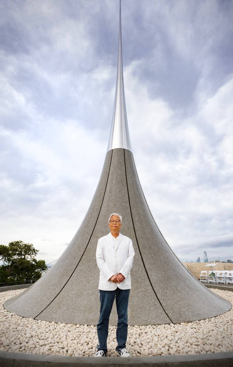 A man in a white coat and jeans stands in front of a large sculpture.
