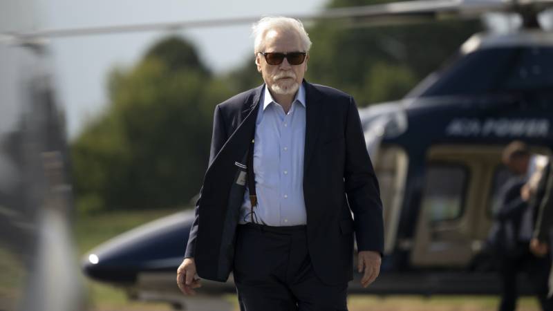 An older man in a suit walks confidently away from a helicopter. He has white hair, a white beard and is wearing sunglasses.