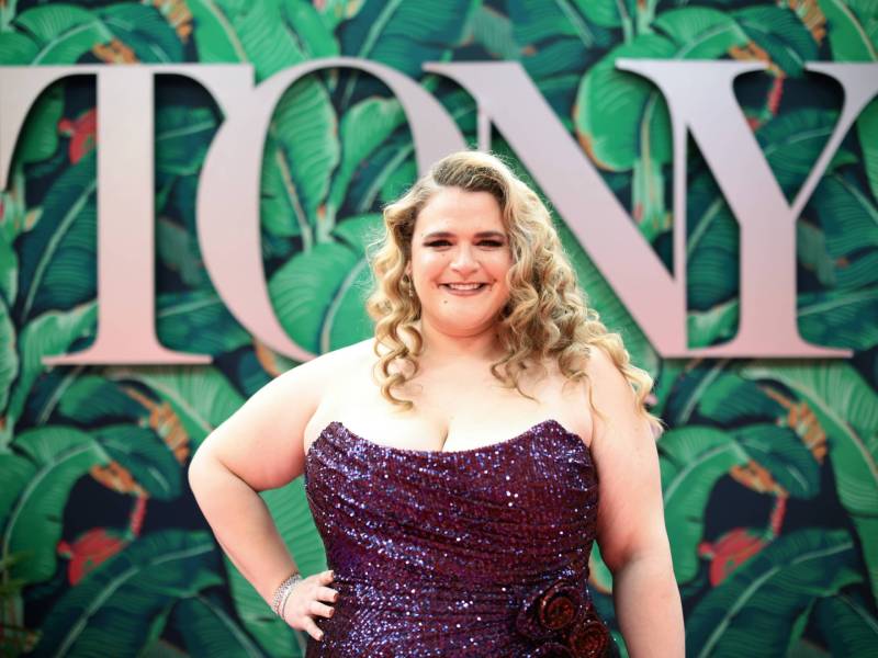 A curvy white blonde woman wearing a purple strapless gown stands in front of a wall with TONY written on it.