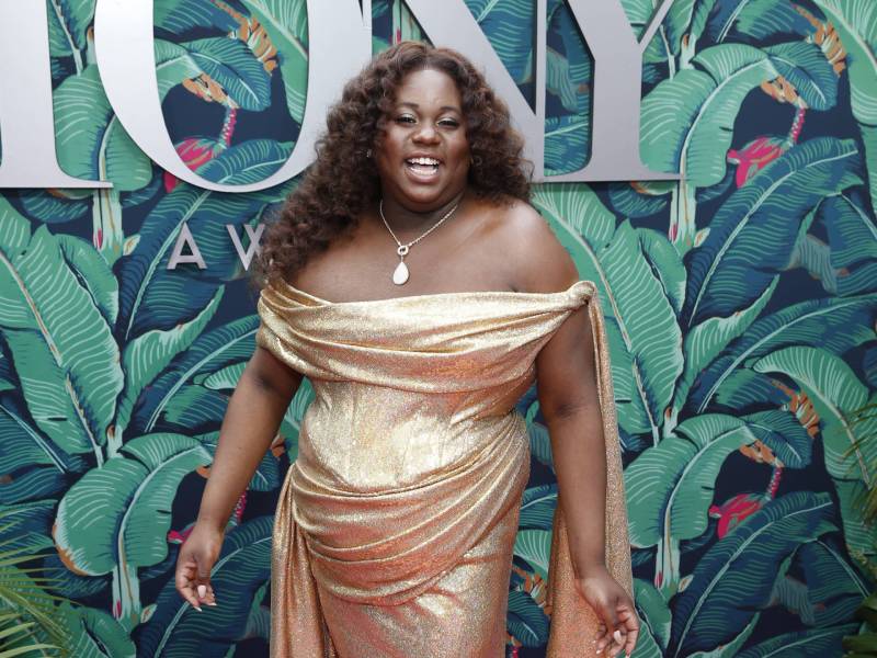 A curvy Black woman in a gold gown smiles and strikes a pose in front of a green wall with TONY written on it.