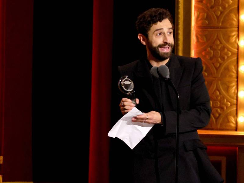 A man with dark curly hair and beard stands on stage holding a trophy and a sheet of paper. He is smiling broadly.