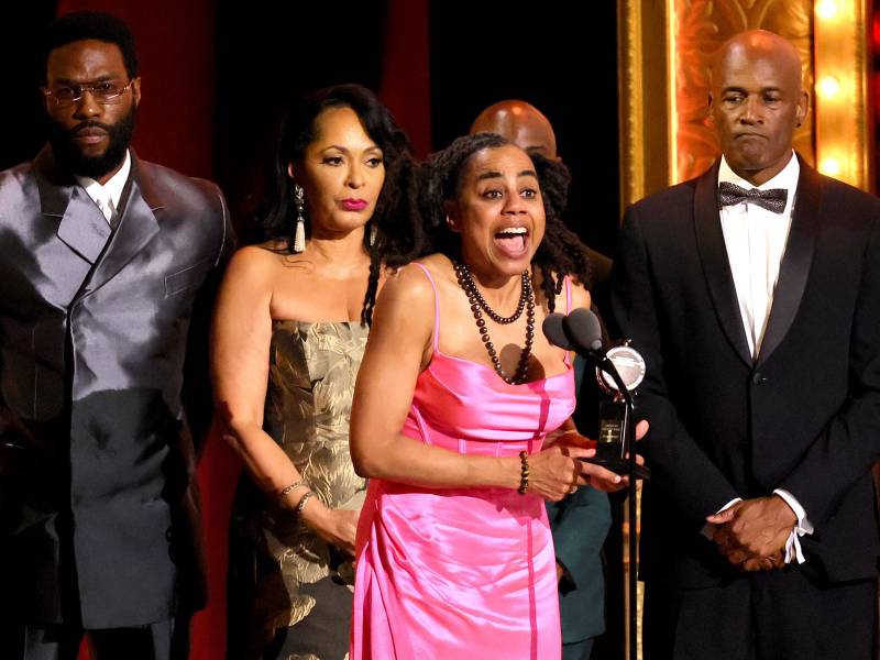 A Black woman in a pink dress gasps with joy while accepting a trophy at a podium. Behind her stand three Black men in suits and a woman wearing a gold gown.