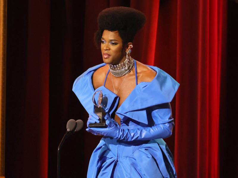 A nonbinary Black person in a powder blue gown and long gloves holds a trophy and leans forward towards a microphone.