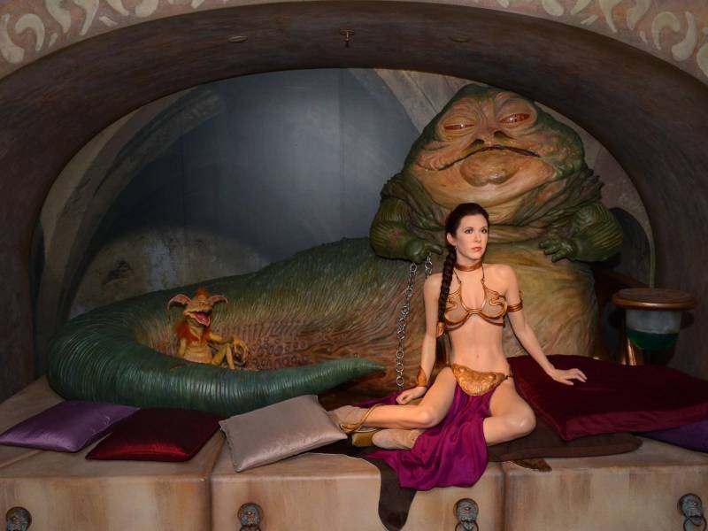 A wax figure of a white woman seated on the ground wearing a gold bikini with purple skirt attachment. Behind her is a large monster that resembles a slug.