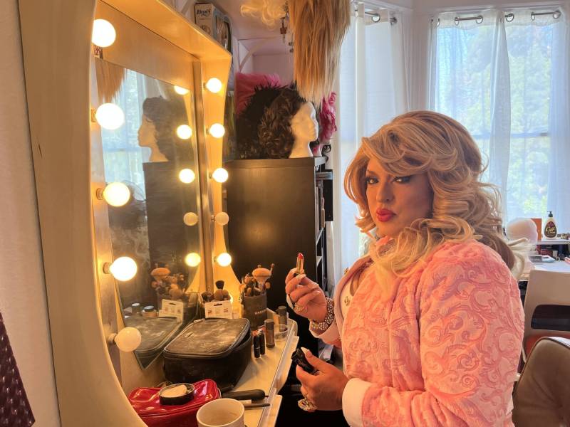 A drag queen in a blond wig sits before a stage makeup mirror, lipstick in hand. She is pouting.