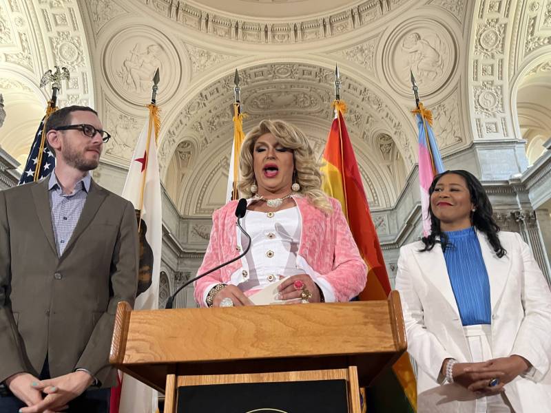 A drag queen in a pink and white suit stands at a podium and speaks, with a Black woman in a white suit to her right, and a white man in a grey suit to her lift. A series of flag poles are visible lined up behind her.