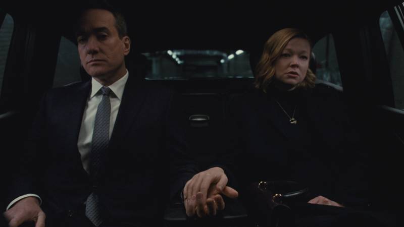 A white man in a suit and a white woman wearing black sit in the back of a car, street lights visible through the back window. Her hand gently rests on his in between them.
