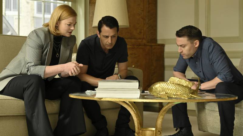 A woman and two men, all white, huddle around a coffee table, listening closely to a message being relayed.