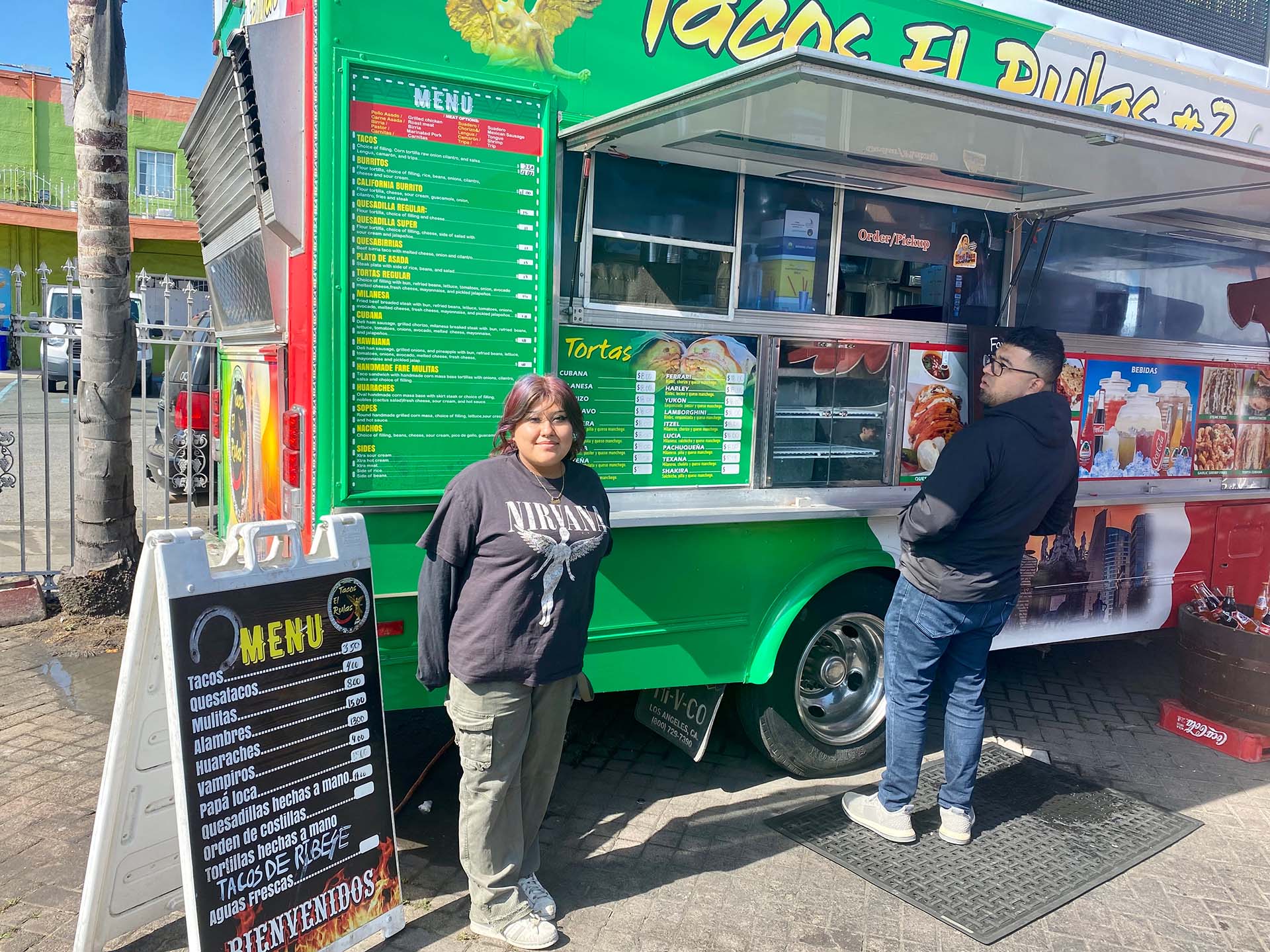 A teenage girl in a black long-sleeved Nirvana T-shirt stands in front of a green taco truck.