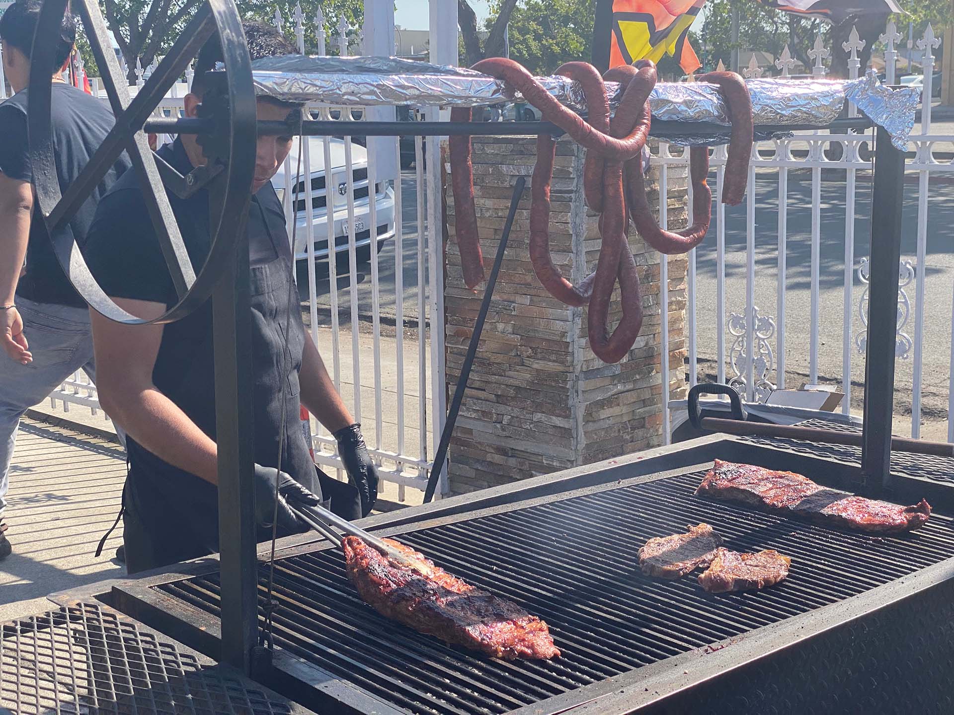 A man in black uses tongs to flip a rack of ribs cooking on the grill. A long chorizo sausage is coiled overhead on the frame of the grill.