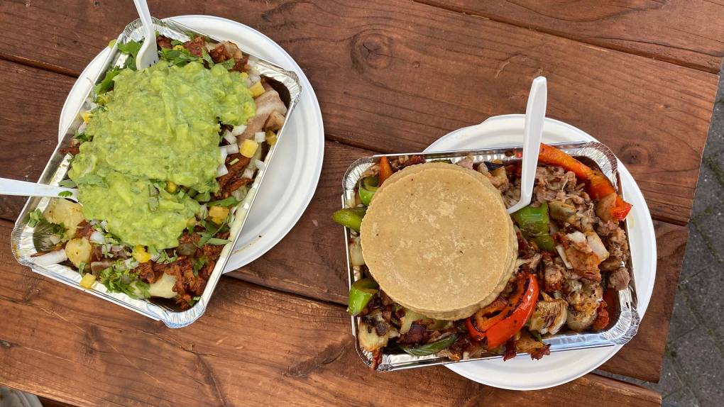 Overhead view of two Mexican dishes served in aluminum takeout trays, on a picnic table. On the left, potatoes are topped with meat, onions and guacamole. On the right is a cheesy mixed grill of onions, peppers and assorted meats, topped with a stack of tortillas.
