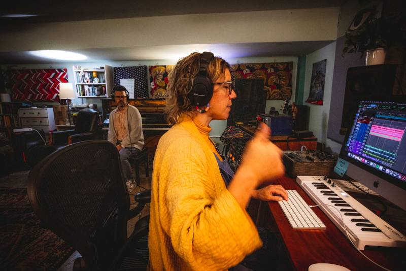 a man and a woman in a music studio, the woman is wearing headphones and sitting at a computer and giving a thumbs up