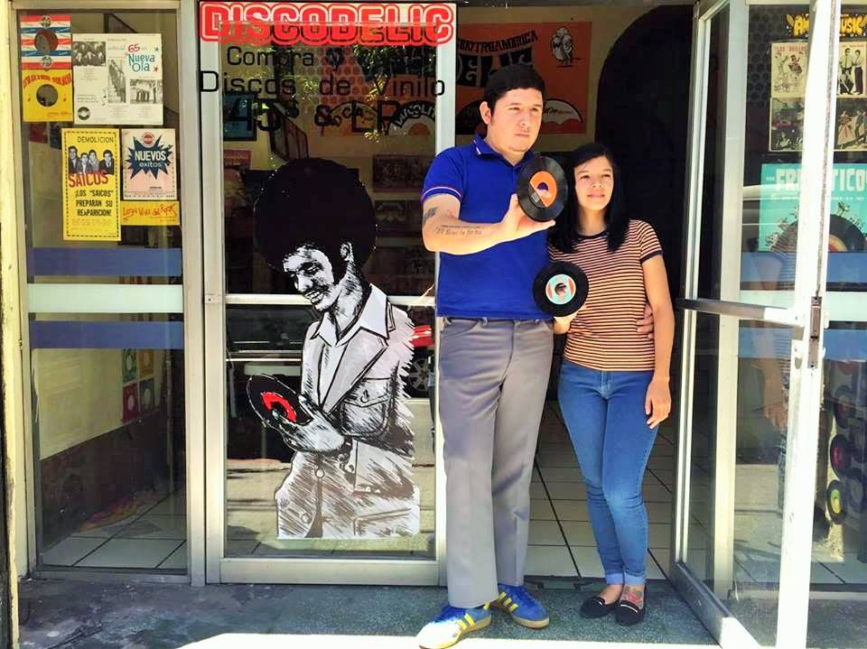 a Latino man and a Latina woman stand together outside the door of a record store they own