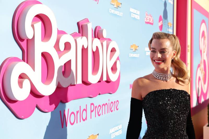 A beautiful blonde woman smiles in the sunshine on a red carpet, wearing a black strapless dress, long gloves and pearl necklace. Behind her is a pink and white Barbie logo.