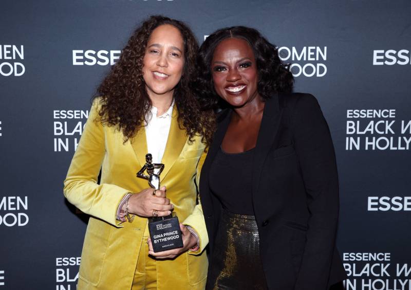 Two Black women with shoulder length wavy hair stand close together, smiling, on a red carpet. The woman on the left wears a gold velvet suit and white shirt and clutches an award statue. The woman on the right wearing a Black suit.