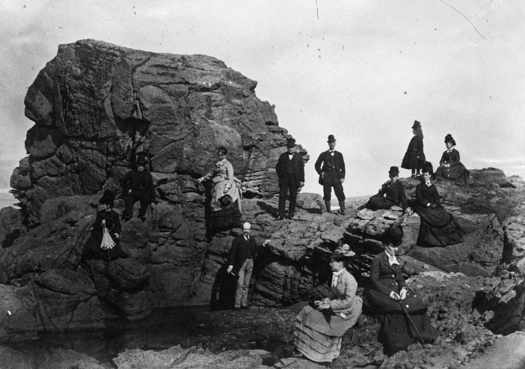 Four men, six women and a child, all dressed in Victorian era clothing sit and stand on a rocky outcrop.
